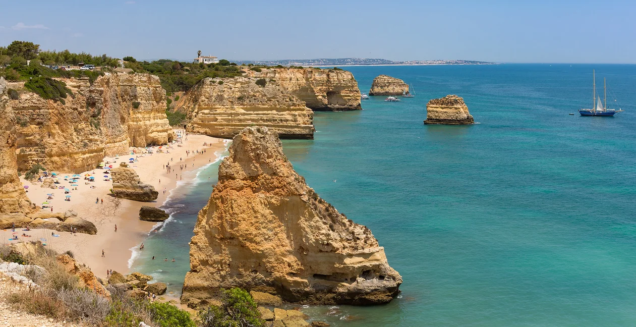 Praia cercada por falésias de arenito com pessoas e guarda-sóis na areia, e embarcações no mar azul-turquesa.