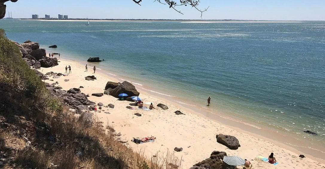 Vista de uma pequena praia com pessoas relaxando e desfrutando do sol, areia clara e rochas, com águas calmas e um veleiro ao fundo.