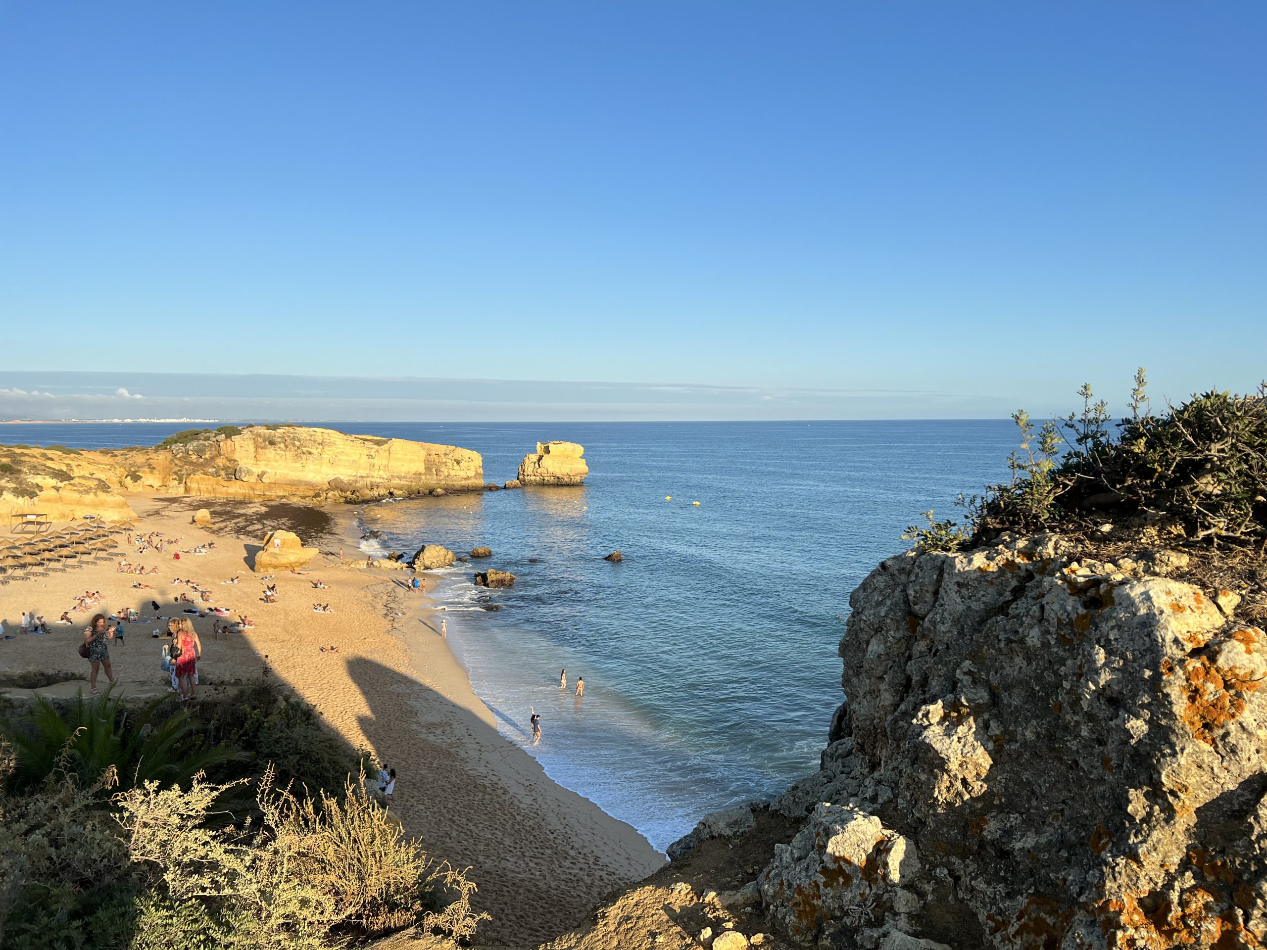 Praia lotada com rochas grandes e o oceano ao fundo sob um céu azul claro.